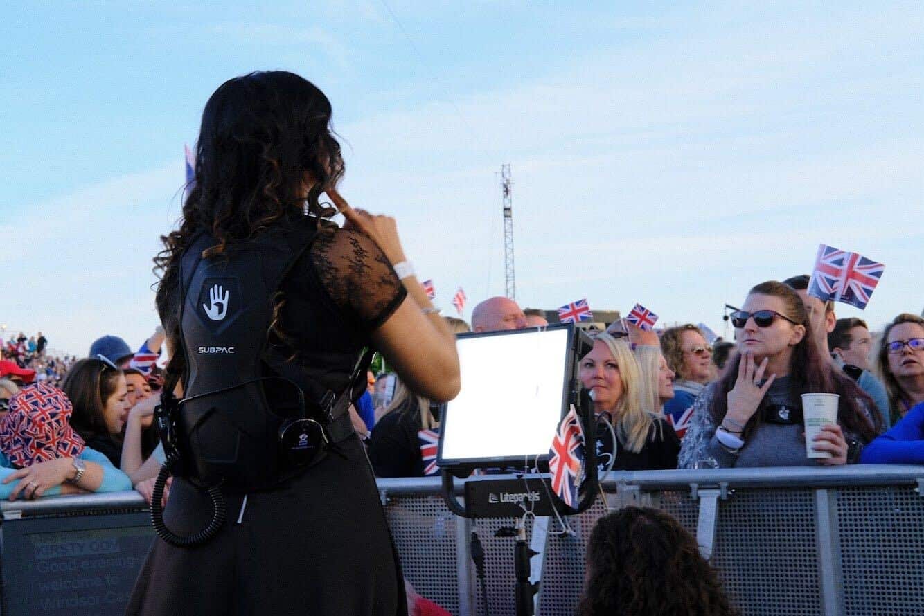 A BSL interpreter, equipped with a SUBPAC device, signs the Coronation Concert performance to a Deaf concertgoer, also wearing a SUBPAC, enhancing their shared musical experience.