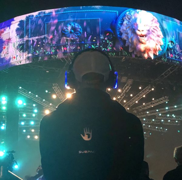 A concert attendee deeply engaged in the Coronation Concert experience feeling the rhythm through the SUBPAC wearable technology, with the vibrant concert stage in the backdrop